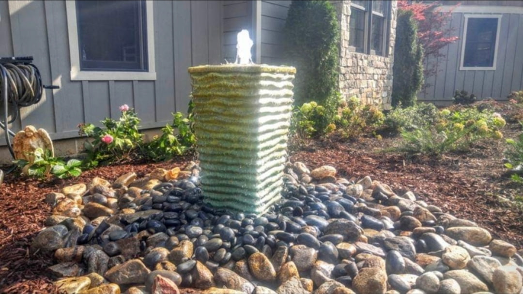 Tall green ribbed column fountain in a black river-rock bed