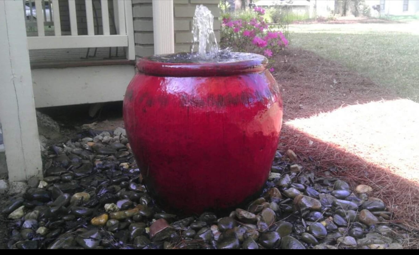 Glossy red jar fountain with a water spout in a river-rock bed